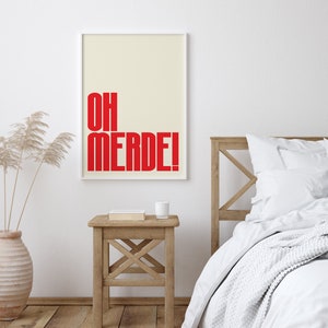 May include: A white framed poster with a cream background and red text that reads "OH MERDE!" hanging on a white wall above a wooden bedside table with a book and a white cup on top. A white bed with a white blanket and a white pillow is in the background.