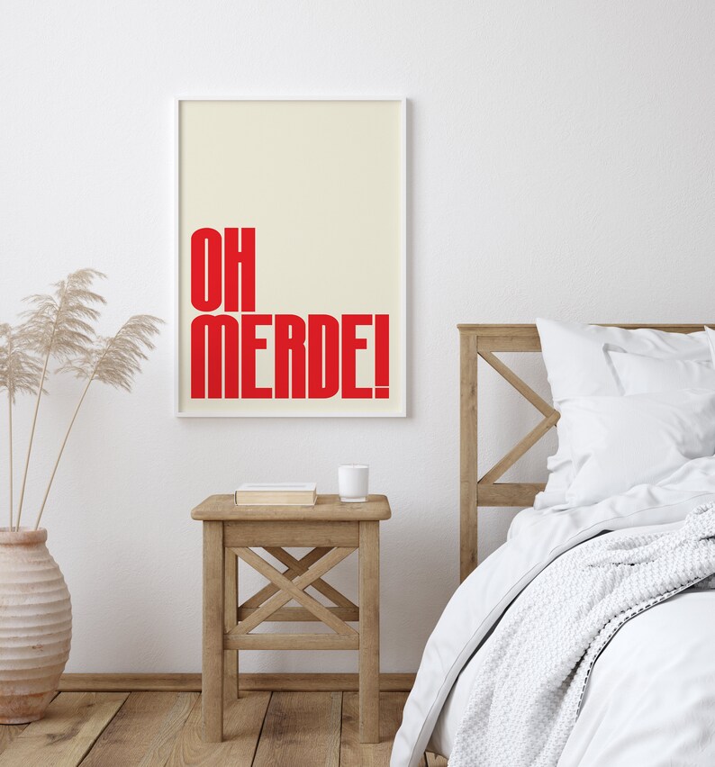 May include: A white framed poster with a cream background and red text that reads "OH MERDE!" hanging on a white wall above a wooden bedside table with a book and a white cup on top. A white bed with a white blanket and a white pillow is in the background.