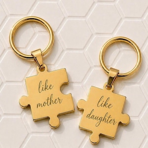 Pode incluir: Dois chaveiros dourados, cada um em forma de peça de quebra-cabeça. Um diz "like mother" e o outro "like daughter". Os chaveiros têm um anel circular na parte superior para prender as chaves. O fundo é um padrão de azulejos hexagonais brancos.