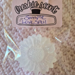 Könnte beinhalten: Ein klarer, blütenförmiger Wachsschmelz in einer transparenten Plastiktüte. Die Tüte hat ein rosa Etikett mit den Worten "Freshie Scent" und "Sample Love Spell". Der Hintergrund ist ein rosa Strickstoff.