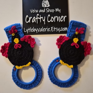 May include: Two handmade crochet towel rings. Each ring is blue with a black chicken design, featuring red comb and wattle, and yellow beak. The sign in the background reads "Crafty Corner" and the Etsy shop URL.