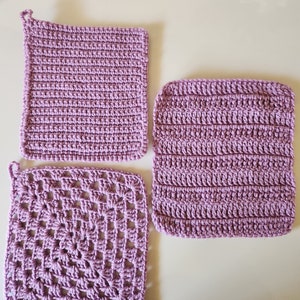 May include: Three purple crocheted dishcloths, one with a simple stitch pattern, one with a striped pattern, and one with a more intricate square pattern.