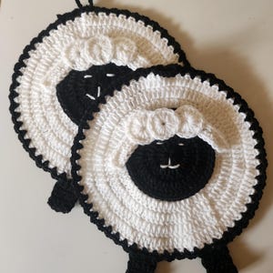 以下が含まれることがあります： 手作りの羊の形をしたかぎ針編みの鍋つかみ2つ。鍋つかみは白で、羊の顔、足、縁取りなど、黒のアクセントが付いています。各鍋つかみには吊り下げ用のループが付いています。羊の顔は、白いステッチのディテールが施された黒い円です。