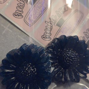 Könnte beinhalten: Zwei dunkelblaue, blütenförmige Artikel im Vordergrund. Die Blumen haben detaillierte Blütenblätter und ein strukturiertes Zentrum. Im Hintergrund befinden sich mehrere rosa Etiketten mit dem Text "Freshie Scents" in Schreibschrift.