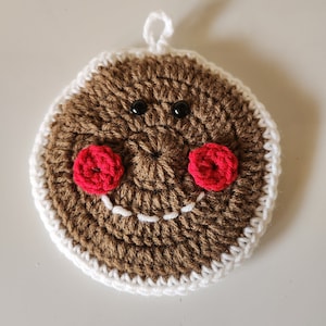 Puede incluir: Un posavasos de hombre de jengibre tejido a ganchillo con una cara sonriente. El posavasos es marrón con un borde blanco y mejillas rojas. Tiene ojos de cuentas negras y una sonrisa cosida blanca. Hay un pequeño lazo blanco en la parte superior para colgar.