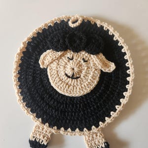 Op de afbeelding: Een handgemaakte, ronde gehaakte onderzetter in de vorm van een schaap. Het schaap heeft een zwarte body, een beige gezicht en poten, en een zwarte, pluizige bovenkant. De onderzetter is ontworpen om oppervlakken te beschermen tegen hete voorwerpen.