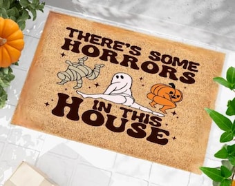 May include: A brown coir doormat with the text "There's some horrors in this house" in white and black lettering. The doormat features a cartoon mummy, ghost, and pumpkin.
