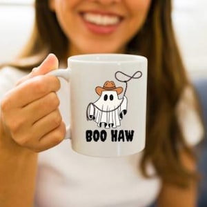 May include: White ceramic coffee mug with a ghost wearing a cowboy hat and holding a lasso. The text "BOO HAW" is printed on the mug.