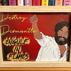May include: A framed print with a light wood frame. The print features a cartoon illustration of a man with a beard and glasses, pointing upwards. Text on the print reads "Destroy, Dismantle, Engulf in Flames" in yellow and white.