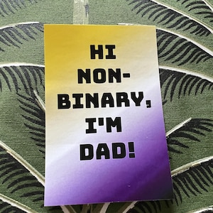 May include: A rectangular card with a yellow, white, and purple gradient. The text "HI NON-BINARY, I'M DAD!" is printed in bold, black capital letters. The card is placed on a green and white patterned surface, resembling a leaf design.