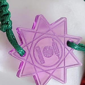 Puede incluir: Primer plano de un dije morado en forma de estrella con un cordón verde. El dije tiene un diseño geométrico con un emblema circular en el centro. El cordón es de material verde trenzado. El dije es probablemente parte de una pulsera o collar.