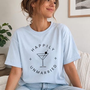 Puede incluir: Una camiseta de manga corta de color azul claro con un gráfico negro de una copa de martini con una aceituna. El texto "HAPPILY UNMARRIED" está impreso en disposición circular alrededor de la copa, junto a pequeños iconos de estrellas.