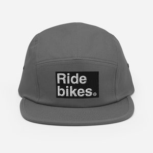 May include: A gray five-panel cap with a flat brim. The cap features the embroidered text "Ride bikes" in white on a black rectangular patch. The cap is made of a durable fabric and has a classic, sporty design.