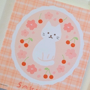 May include: A pink and white sticker with a white cat sitting in the center of a circle. The circle is surrounded by pink cherry blossoms and red cherries. The sticker has a checkered background.