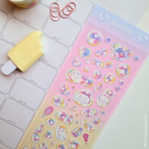 May include: A sheet of bubble-themed stickers with various shapes and designs, including hearts and cartoon characters, in a gradient of pink, yellow, and blue. A yellow popsicle-shaped eraser and pink heart-shaped paper clips are also present.