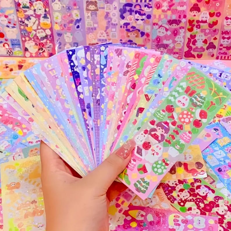 Op de afbeelding: Een hand die een waaier van kleurrijke stickerbladen vasthoudt. De stickers bevatten diverse ontwerpen, waaronder kerstthema-illustraties, sterren en andere schattige patronen. Het kleurenpalet omvat pasteltinten zoals roze, paars en groen.