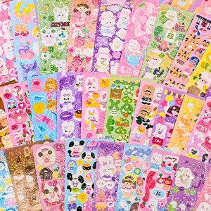 Op de afbeelding: Een verzameling kleurrijke sticker vellen met diverse ontwerpen. De stickers tonen schattige illustraties van dieren, voedsel en decoratieve elementen. Het kleurenpalet omvat roze, paars, groen en geel, met glitter accenten.