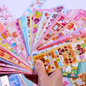 Op de afbeelding: Een hand houdt een waaier van kleurrijke stickerbladen vast. De stickers hebben verschillende ontwerpen, waaronder cartoon dieren, voedsel en decoratieve patronen. Het kleurenpalet omvat roze, blauw, geel en groen, wat een speelse en grillige uitstraling cre&euml;ert.