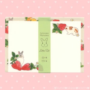May include: A stationery set featuring a bunny and strawberry design. The set includes white paper with illustrations of bunnies and strawberries. The packaging is green and has the text "Schinako Moriyama Letter Set". The background is pink with heart shapes.