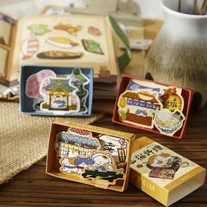 May include: Four small boxes with colorful stickers of Japanese food and cats. The boxes are made of cardboard and have a brown and white design. The stickers are made of paper and have a glossy finish. The stickers feature images of sushi, ramen, and other Japanese food, as well as cats in various poses. The boxes are arranged in a row on a wooden table.