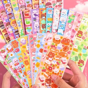 Op de afbeelding: Een verzameling kleurrijke sticker vellen met verschillende schattige illustraties. Ontwerpen omvatten beren, konijntjes, cakes en andere voedingsmiddelen. De stickers zijn in roze, blauw, geel en groen, met een kawaii-esthetiek.