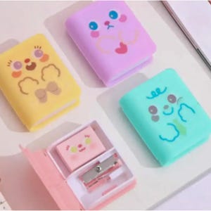 May include: Four colorful pencil sharpeners shaped like books with cartoon animal faces. Each sharpener has a different color and animal design. The sharpeners are on a white surface.