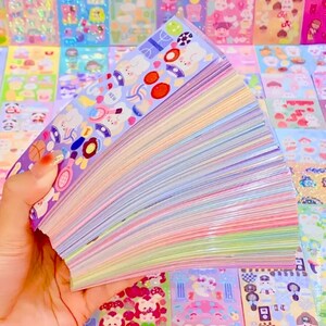 Op de afbeelding: Een hand houdt een stapel kleurrijke stickers met verschillende ontwerpen vast. De stickers bevatten illustraties van sportartikelen, eten en stripfiguren. De randen van de stickers tonen een regenboog van pastelkleuren.