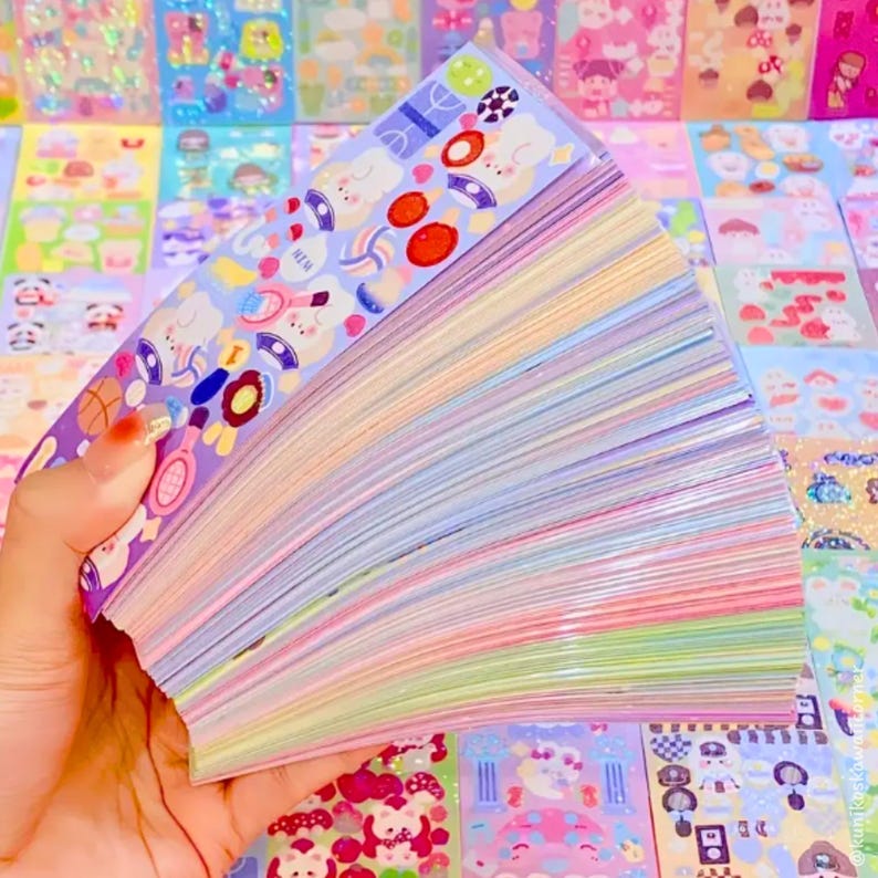 Op de afbeelding: Een hand houdt een stapel kleurrijke stickers met verschillende ontwerpen vast. De stickers bevatten illustraties van sportartikelen, eten en stripfiguren. De randen van de stickers tonen een regenboog van pastelkleuren.