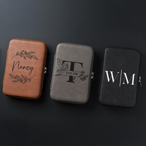 Three rectangular leatherette tech cases in brown, grey, and black lie side by side on a dark surface, each featuring a different customized engraving with names and initials.