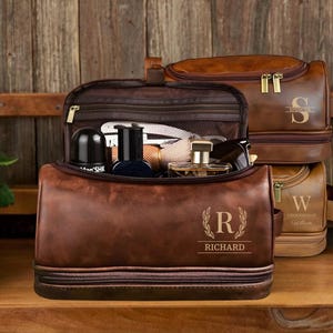 May include: Two brown leather travel bags with zipper closures. The larger bag has a monogrammed "S" and the smaller bag has a monogrammed "W". The smaller bag has the text "Groomsmans Gift" on it. The larger bag is open and has a brown mesh lining. The bag is filled with toiletries and other travel essentials. The bag has a monogrammed "R" with a laurel wreath design and the name "Richard" underneath.