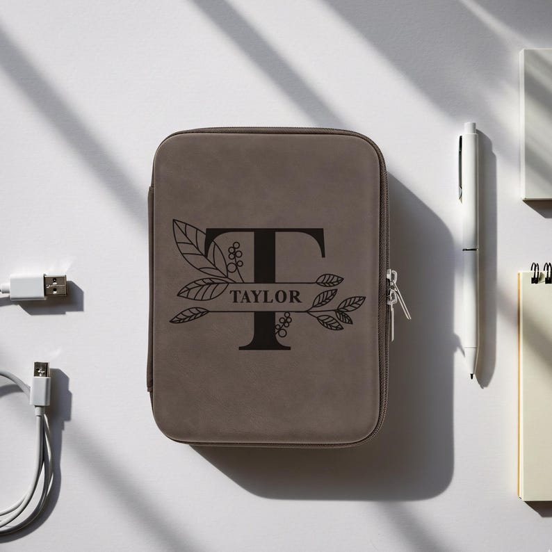 Grey leatherette tech case engraved with a large letter T, botanical branches, and the name Taylor, placed on a white surface with a pen, notebook, and USB cables nearby.