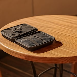 May include: Two open black zippered cases on a wooden table. One case is open, revealing a mesh interior and straps. The other case is closed with a woven strap design. The table is round with a light brown finish.