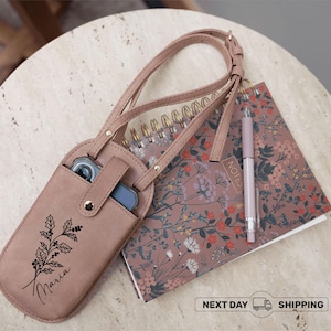 May include: A blush-colored phone crossbody bag with a floral design and the name "Maria" printed on it. The bag has a strap and a snap closure. A floral notebook and pen are next to the bag.