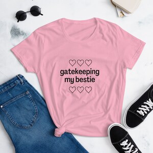 May include: A light pink t-shirt with the text "gatekeeping my bestie" and three heart symbols above and below the text. The shirt is paired with blue jeans and black sneakers.