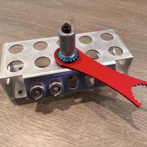 May include: A metal tool set with a red wrench, a silver cylinder, and a blue gear. The set is placed on a silver metal holder with multiple circular openings. The tools are designed for mechanical or engineering applications.