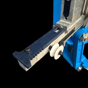 May include: Close-up of a blue and silver metal machine component. The component has a blue rubberized edge with a ridged pattern, two white wheels, and silver metal parts. The background is black and blue.