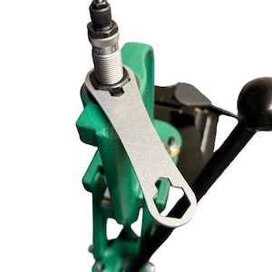 Puede incluir: Una prensa de recarga con una llave plateada sujeta a un marco verde. La prensa tiene un mango negro y una varilla roscada. La llave tiene una abertura hexagonal.