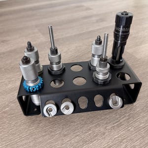 Puede incluir: Un organizador de herramientas de metal negro que contiene varias herramientas de recarga. Las herramientas incluyen componentes plateados y negros, algunos con detalles azules. Una herramienta está etiquetada como "MatchMaster".