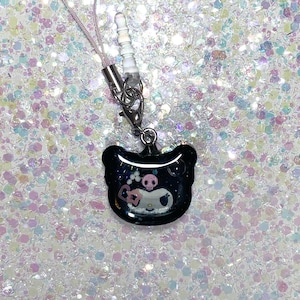 May include: A black phone charm with a bear-shaped pendant. The pendant features a cartoon design with a skull, a ghost, and a bow. The charm has a pink cord and a silver-colored metal clasp and connector.