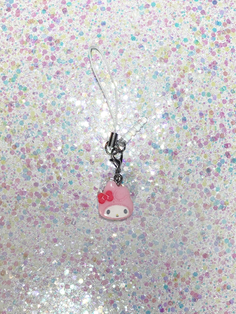 Puede incluir: Un charm de tel&eacute;fono rosa de My Melody con un lazo rojo y detalles blancos. El charm est&aacute; unido a un cierre de metal plateado y un cord&oacute;n blanco. El fondo es una superficie brillante y con purpurina.