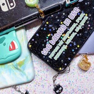 Puede incluir: Funda de tel&eacute;fono negra con un patr&oacute;n de estrellas brillantes y el texto "the babies in use". Incluye colgantes de un rollo de sushi, un oso y un gato negro. Tambi&eacute;n incluye una Nintendo Switch y una funda Bulbasaur.