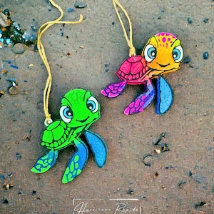 May include: Two colorful wooden turtle ornaments with string hangers. One turtle is green with blue accents and the other is pink, orange, and purple.
