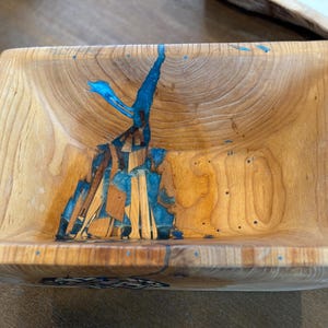 May include: A rectangular wooden bowl with a natural wood grain finish. The bowl features a striking blue resin inlay that adds a pop of color and visual interest. The bowl is handcrafted and has a unique design.