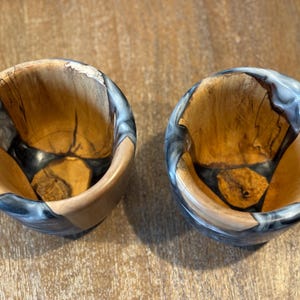 Puede incluir: Dos cuencos de madera con interior de vetas de madera natural y exterior de resina gris oscuro y blanco. Los cuencos son redondos y tienen un acabado liso. Los cuencos son probablemente para servir alimentos o como artículos decorativos.