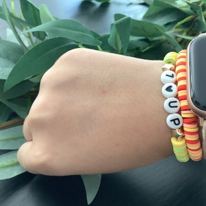 Könnte beinhalten: Ein Armband mit weißen Perlen, die "I ♥ UP" buchstabieren, und zwei bunte Perlenarmbänder. Die Armbänder werden an einem Handgelenk mit einer goldenen Apple Watch getragen.
