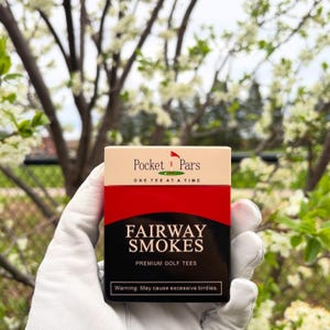 May include: A box of "Pocket Pars" golf tees. The box has a cream-coloured top with the text "Pocket Pars" and "ONE TEE AT A TIME". The bottom half is red and black with the words "FAIRWAY SMOKES" and "PREMIUM GOLF TEES".