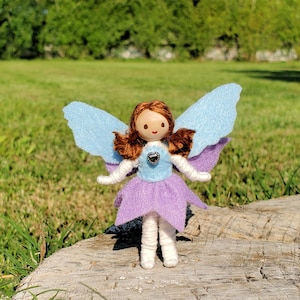 May include: A handmade doll with brown hair, a blue and purple dress, and blue felt wings. The doll is standing on a piece of wood.