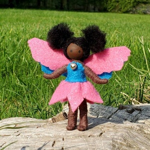 May include: A handmade fairy doll with a wooden head, black yarn hair, and pink and blue felt wings and dress. The doll has brown yarn arms and legs and is standing on a piece of wood.