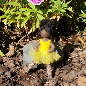 May include: A handmade doll with a dark complexion, black hair, and a yellow tutu. The doll is posed as a ballerina, with wrapped yarn legs and arms. The doll is sitting on a gray rock, with a background of green foliage and a pink and white flower.