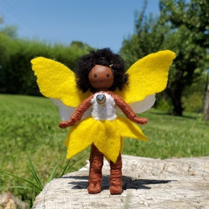 May include: A handmade fairy doll with a wooden head, black hair, and a brown wrapped body. The fairy has yellow and white felt wings, a yellow skirt, and a white top with a blue gem. The doll is standing on a wooden surface.